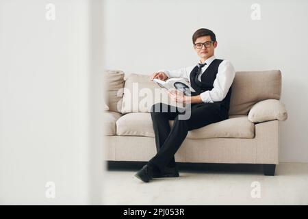 Ein junger Mann in formeller Kleidung sitzt auf dem Sofa drinnen an einer weißen Wand Stockfoto