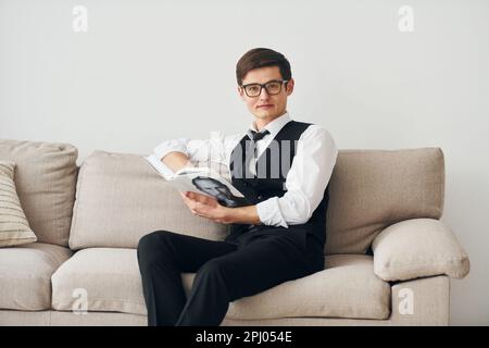 Ein junger Mann in formeller Kleidung sitzt auf dem Sofa drinnen an einer weißen Wand Stockfoto