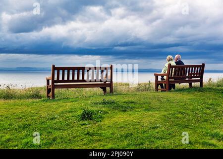 Ältere Ehepaare, die auf einer Holzbank sitzen und den Blick auf Firth of Forth genießen, wolkigen Himmel, North Berwick, East Lothian, Schottland, Vereinigtes Königreich Stockfoto