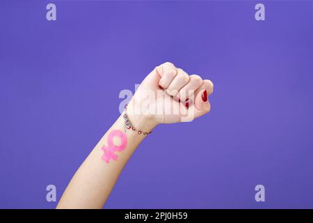 Nahaufnahme der Frauenfaust mit dem feministischen Symbol auf einem isolierten violetten Samthintergrund Stockfoto