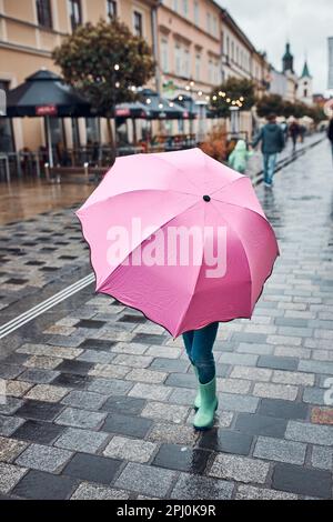 Kleines Mädchen versteckt sich hinter großen rosa Regenschirm zu Fuß in einer Innenstadt an regnerischen düsteren Herbsttag Stockfoto