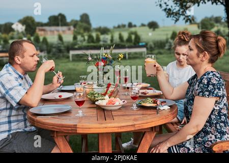 Familie mit einer Mahlzeit vom Grill während des Sommer Picknick im Freien Abendessen in einem Hausgarten. Nahaufnahme von Leuten, die an einem Tisch mit Essen und Geschirr sitzen Stockfoto