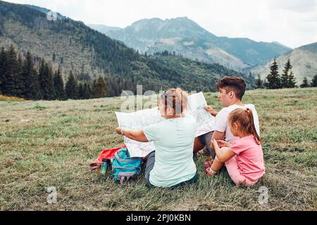 Familie, die eine Karte mit Bergversuchen durchsucht, auf dem Rasen sitzt und den Sommertag während des Urlaubs in den Bergen genießt. Menschen verbringen aktiv Zeit im Freien Stockfoto