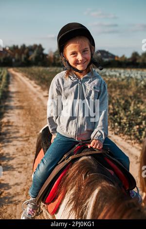 Kleines lächelndes Mädchen, das Reiten lernt. 5-6 Jahre alter Pferdesport im Helm mit Spaß Reiten ein Pferd Stockfoto