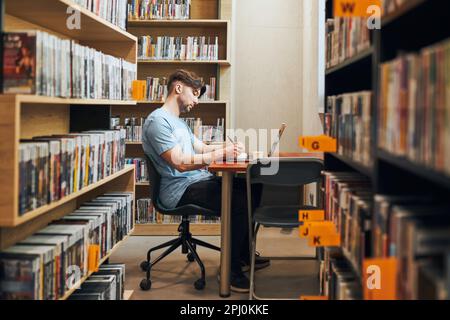 Schüler lernen in der Bibliothek. Junger Mann, der sich auf den Test am Laptop vorbereitet. Mann hört sich Online-Kurs an. Fokussierter Student, der für College-Prüfungen studiert. Verwenden Von Stockfoto