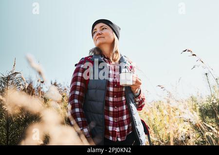 Frau macht Pause und entspannt sich mit einer Tasse Kaffee während der Sommerreise. Eine Frau, die auf der Spur steht und wegschaut. Frau mit Rucksack, die durch Ta wandert Stockfoto