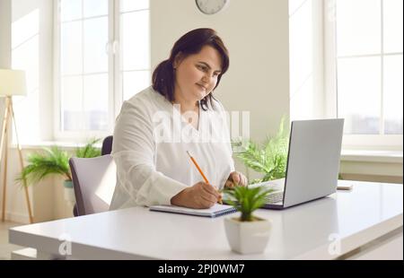 Übergewichtige braune junge Frau, die im modernen Büro ein Notebook benutzt Stockfoto
