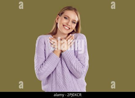 Lächelnde Blondine in lila Pullover, die Hände auf Khaki-Hintergrund an der Brust oder am Herzen hält. Stockfoto