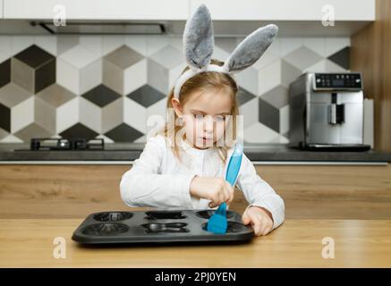 Ernstes Kind kleines Mädchen in Hasenohren, das in der modernen Holzküche zu Hause die Backform mit einer blauen Silikonbürste mit Olivenöl schmiert. Osterkonzept, Stockfoto