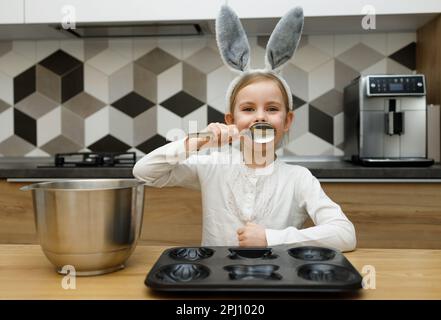 Ein Kind mit Hasenohren lacht und lächelt, bedeckt ihren Mund mit Löffel, kocht und backt Kekse, Muffins, hat Spaß in der Küche. Frohe Ostern, Stockfoto