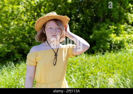 Ein asiatisches Teenager-Mädchen in einem gelben Kleid, eine Brille richtet ihren Hut im Park auf ihren Kopf. Serious Girl mit Hut vor dem Hintergrund von grünem Urlaub Stockfoto