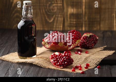 Reifer Granatapfelbruch, verstreute große Körner, frisch gepresster Granatapfelsaft in einer transparenten Flasche auf Holzhintergrund. Stockfoto