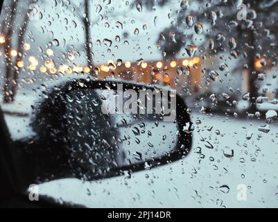 Nahaufnahme Von Wassertropfen Am Seitenfenster Des Autos Während Des Schneefalls Im Winter, Vor Dem Hintergrund Des Rückspiegels Und Der Straßenbeleuchtung Von Bokeh. Stockfoto