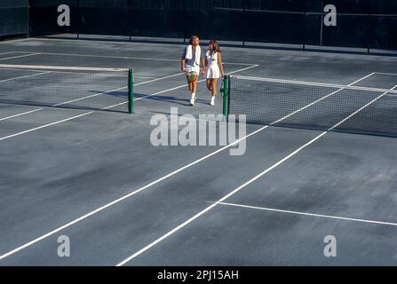 EIN JUNGES PAAR, DAS ZUSAMMEN AUF DEM SANDTENNISPLATZ SPAZIERT Stockfoto