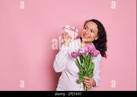 Glückliche schwangere Frau mit welligen Haaren, mit einem Strauß Tulpen, lächelnd, schüttelnd Geschenkbox, erraten, was darin ist Stockfoto