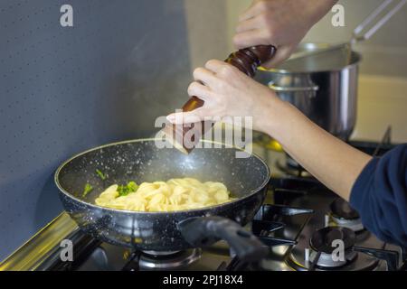 Bild einer Frauenhände, die frisch gekochte Pasta in einer Pfanne mit einer Pfeffermühle würzen Italienische kulinarische Tradition. Stockfoto