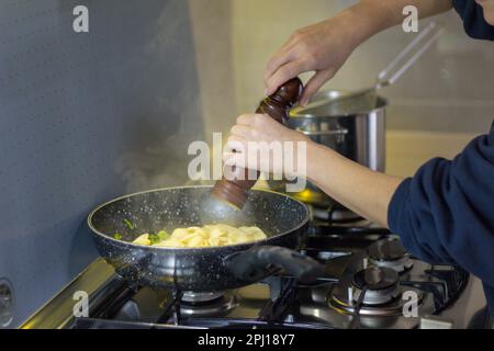 Bild einer Frauenhände, die frisch gekochte Pasta in einer Pfanne mit einer Pfeffermühle würzen Italienische kulinarische Tradition. Stockfoto