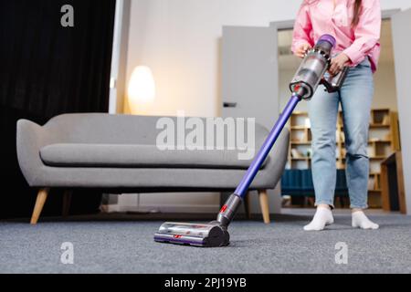 Die junge Frau reinigt den Teppich mit einem kabellosen Staubsauger. Moderne, einfache Reinigung. Stockfoto