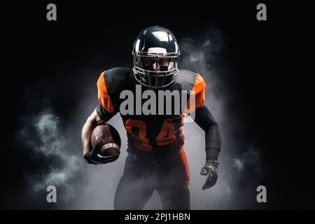 Amerikanischer Fußballspieler auf dunklem Hintergrund in Rauch in schwarz-orangefarbener Ausrüstung Stockfoto