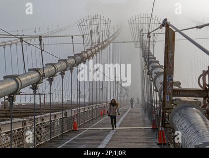 Die berühmten gotischen Türme der Brooklyn Bridge sind von einem Januar-Nebel umgeben; nur wenige Fußgänger trotzen dem kalten und windigen Fußweg. Stockfoto