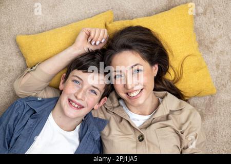 Porträt einer glücklichen Frau mit Sohn, Mutter und Freund, die lächeln und auf die Kamera schauen, zusammen auf Teppichen auf dem Boden zu Hause im Wohnzimmer liegen und Spaß zusammen haben. Stockfoto