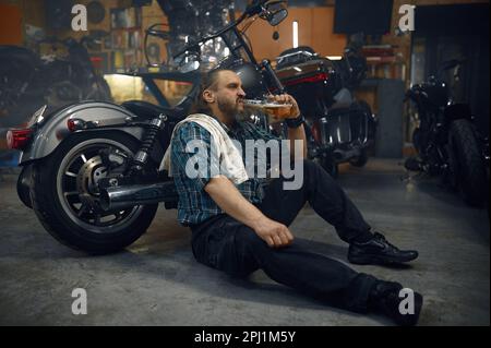 Müder männlicher Biker trinkt Bier aus der Flasche nach harten Reparaturarbeiten in der Garage Stockfoto