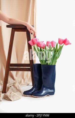 Ein Strauß rosa Tulpen in einem blauen Gummistiefel. Die Hand des Mädchens liegt auf einem Hochstuhl in Beige neben einem Strauß Tulpen in einer blauen Gummistiefel-Vase Stockfoto