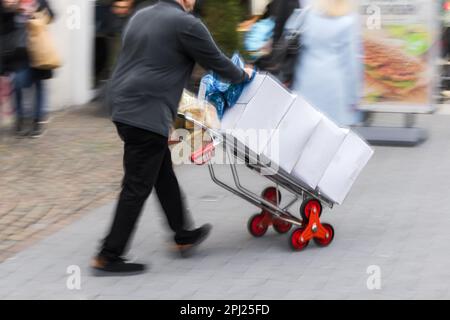 Bild mit Kamera, Zoom-Effekt eines Mannes, der einen Handwagen mit Paketen durch die Fußgängerzone in einer Stadt schiebt Stockfoto