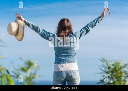 Rückansicht des Glücks Hipster weibliche erhobene Hände genießen Urlaub, stehen auf dem Hintergrund des blauen wolkenlosen Himmels. Eine nicht wiedererkennbare Frau Stockfoto