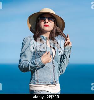 Porträt einer braunen Frau in Denimjacke, Jeans, Strohhut auf dem Kopf und Sonnenbrille, die in die Kamera schaut. Hipster Erwachsene Frau im Stehen Stockfoto