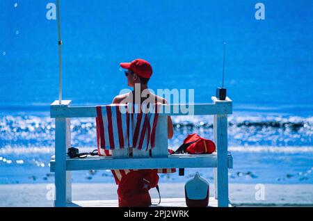 Rettungsschwimmer mit roter Mütze und Sonnenbrille sowie rot-weiß gestreiftem Handtuch sitzen am Sommertag auf seinem Sitzplatz mit Blick auf York Harbor Beach Stockfoto