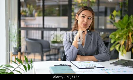 Selbstbewusste und umsichtige asiatische Geschäftsfrau, die über eine Geschäftsstrategie für eine neue Kampagne nachdenkt und an ihrem Schreibtisch arbeitet Stockfoto