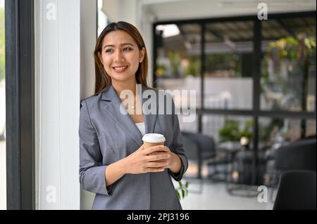 Eine wunderschöne und lächelnde asiatische Geschäftsfrau oder Chefin im formellen Business-Anzug steht am Fenster, hält eine Tasse Kaffee zum Mitnehmen und einen Tag Stockfoto