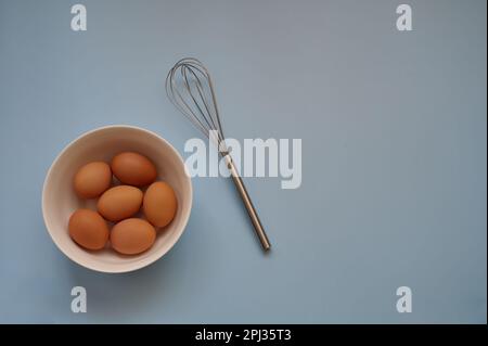 Eier in weißer Schüssel und Schneebesen auf blauem Hintergrund - Draufsicht Stockfoto