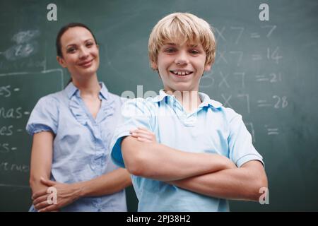 Er ist ihr Starschüler. Ein glücklicher Junge, der seine Arme kreuzte, während sein Lehrer stolz im Hintergrund steht. Stockfoto