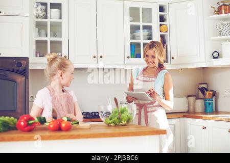 Sie leisten großartige Arbeit, wenn Sie dieses Rezept befolgen. Ein kleines Mädchen, das in der Küche einen Salat macht, während ihre Mutter in der Nähe mit einem digitalen Tablet steht. Stockfoto