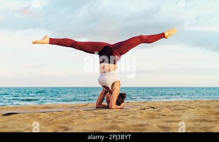 Eine junge weiße Frau in Sportbekleidung übt bei Sonnenuntergang Yoga auf einer natürlichen Matte am Strand und führt eine geteilte Kopfständerhaltung mit dem Meer und Horiz auf Stockfoto