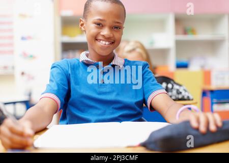 Ich bin bereit, meinen Horizont zu erweitern. Ein kleiner Winkel von einem glücklichen jungen afroamerikanischen Schüler, der auf seinem Schreibtisch sitzt und dich anlächelt. Stockfoto