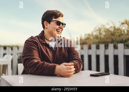 Lächelnde, nicht binäre Person mit Sonnenbrille in einem Café im Freien, die wegschaut. Trägt braune Samtjacke, blauer Himmelshintergrund. Stockfoto