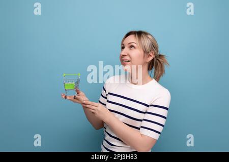 Ein blondes, blondes, attraktives Mädchen in einer lässigen Jacke denkt darüber nach, einen Einkaufswagen in der Hand zu halten, auf blauem Hintergrund Stockfoto