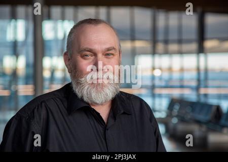 Porträt eines bärtigen Mannes mittleren Alters. Glücklicher älterer Mann, 50 55 60 Jahre alt, mit grauem Bart am Flughafen, Büro, im Innenbereich, der in die Kamera schaut Stockfoto