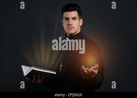 Priester in Cassock hält Bibel mit heiligem Licht auf schwarzem Hintergrund Stockfoto