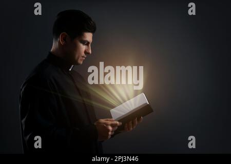 Der Priester hält die Bibel mit heiligem Licht auf schwarzem Hintergrund Stockfoto