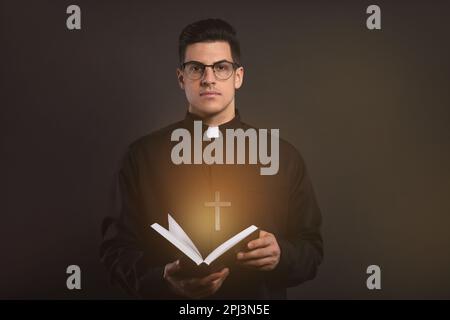 Priester in Cassock hält Bibel mit heiligem Licht auf schwarzem Hintergrund Stockfoto