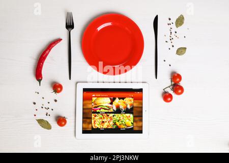 Modernes Tablet mit offener Seite für Online-Lebensmittelbestellung, verstreutes Gemüse, Gewürze, Teller und Besteck auf weißem Holztisch, flach liegend. Konzept von del Stockfoto