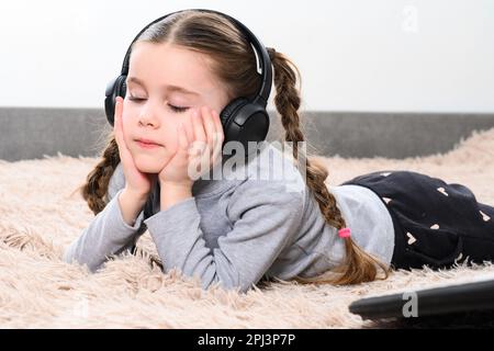 Ein kleines Mädchen liegt auf dem Sofa und hört Musik über Kopfhörer, ein Porträt eines Kindes mit Kopfhörern. Stockfoto