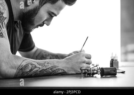 Ein professioneller Tattoo-Künstler, der im Innenbereich am Tisch skizziert. Schwarzweißfotografie Stockfoto