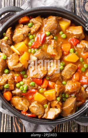 Rindereintopf mit Kartoffeln, Karotten, grünen Erbsen und Gewürzen in einer Pfanne auf einem Holztisch. Vertikal Stockfoto