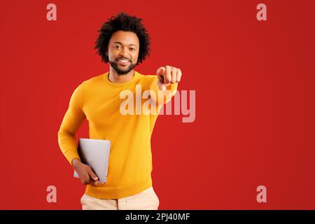 Emotionaler Mann zeigt auf Sie und hält den Laptop, während er mit einem fröhlichen Lächeln in die Kamera schaut. Innenstudio, isoliert auf rotem Hintergrund Stockfoto
