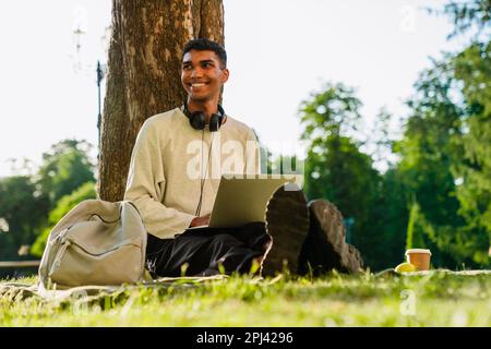 Junger, fröhlicher afrikanischer Mann in Freizeitkleidung, der an einem Laptop arbeitet, während er draußen im Green Park sitzt Stockfoto